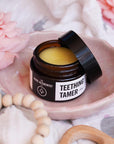 Product shot of Teething Tamer 30g glass jar resting on pink and wooden toned background.